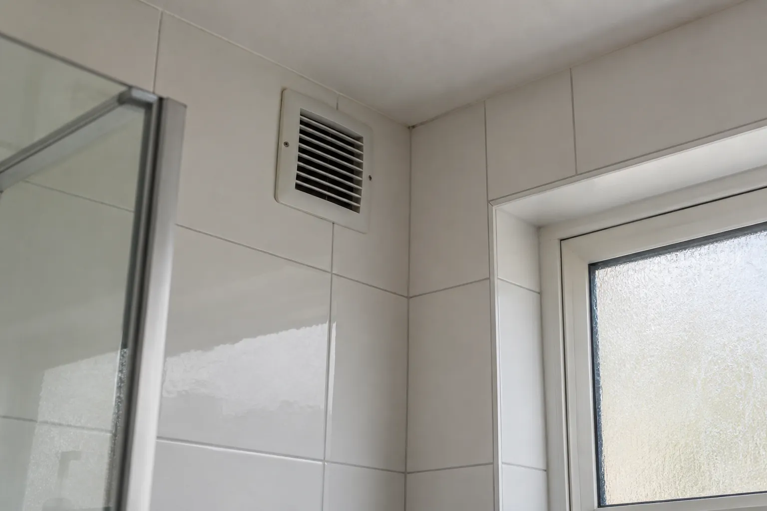 Wit ventilatierooster hoog in een betegelde badkamerwand, naast een douchewand en een raam met matglas — illustratief beeld
