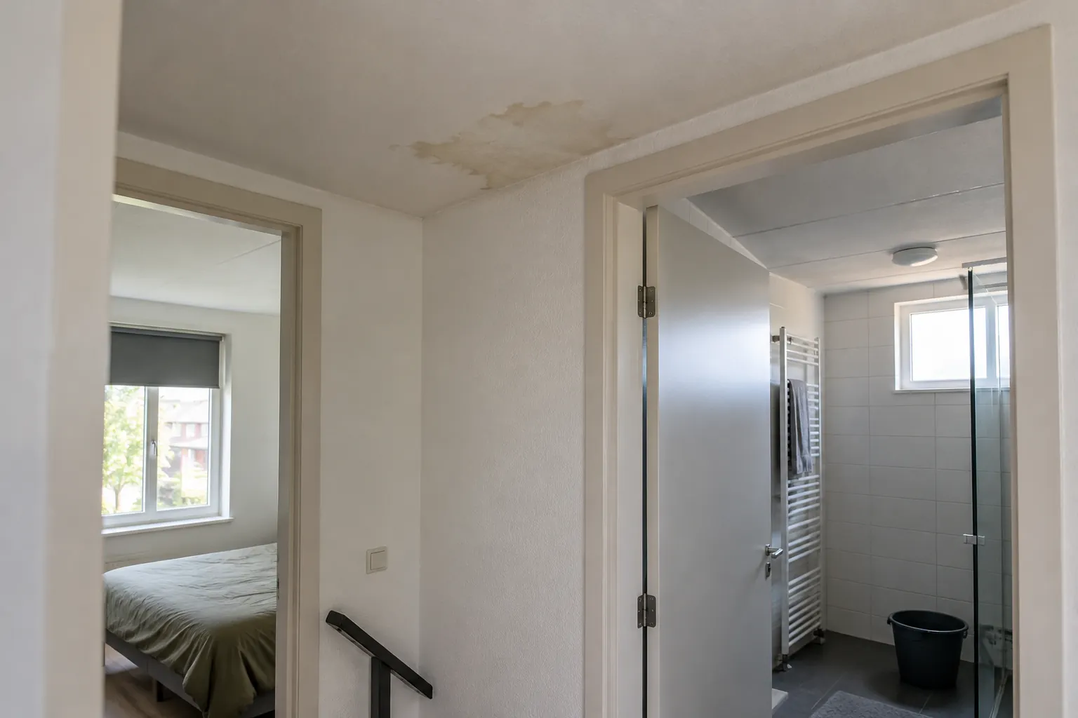 Overloop met een vochtvlek op het plafond, een open badkamerdeur met handdoekradiator en een emmer op de tegelvloer — illustratief beeld