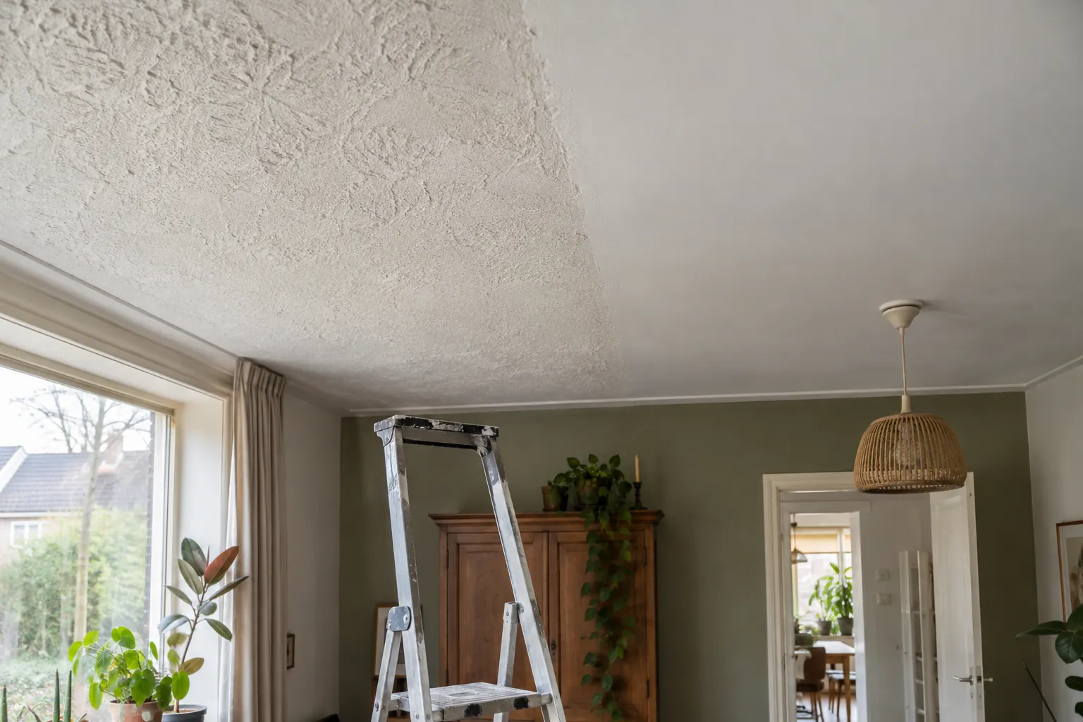 Plafond waarvan een deel grof gestructureerd is en een deel glad wit, met een trapladder eronder in een woonkamer — illustratief beeld