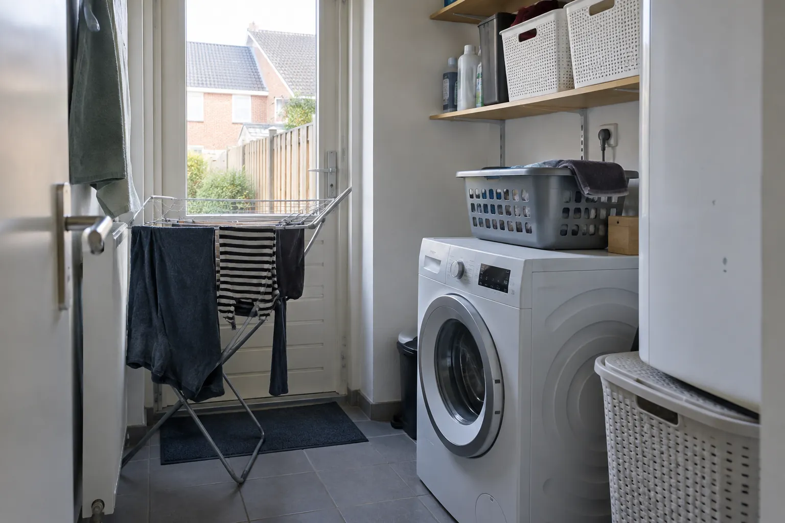 Smalle wasruimte met een wasmachine, een wasmand erop, planken met manden, een droogrek met kleding en een tuindeur — illustratief beeld