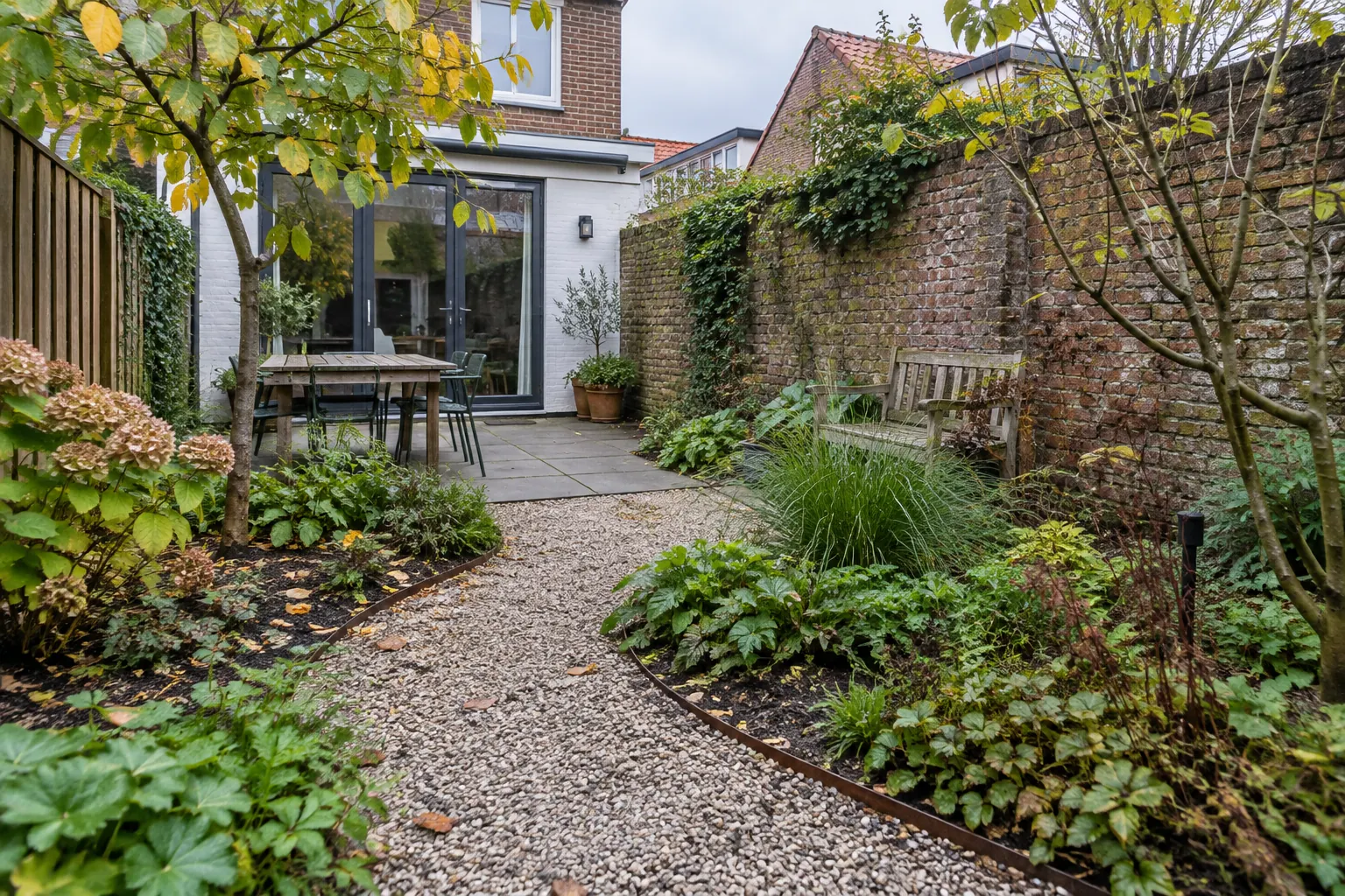 Gebogen grindpad met metalen randen tussen borders in een smalle achtertuin, met terras en eettafel bij het huis — illustratief beeld