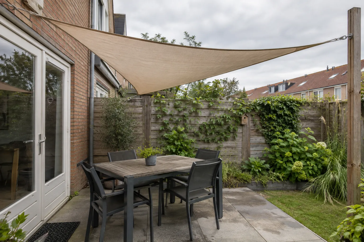 Driehoekig zandkleurig schaduwdoek gespannen tussen gevel en een houten paal boven een tuintafel