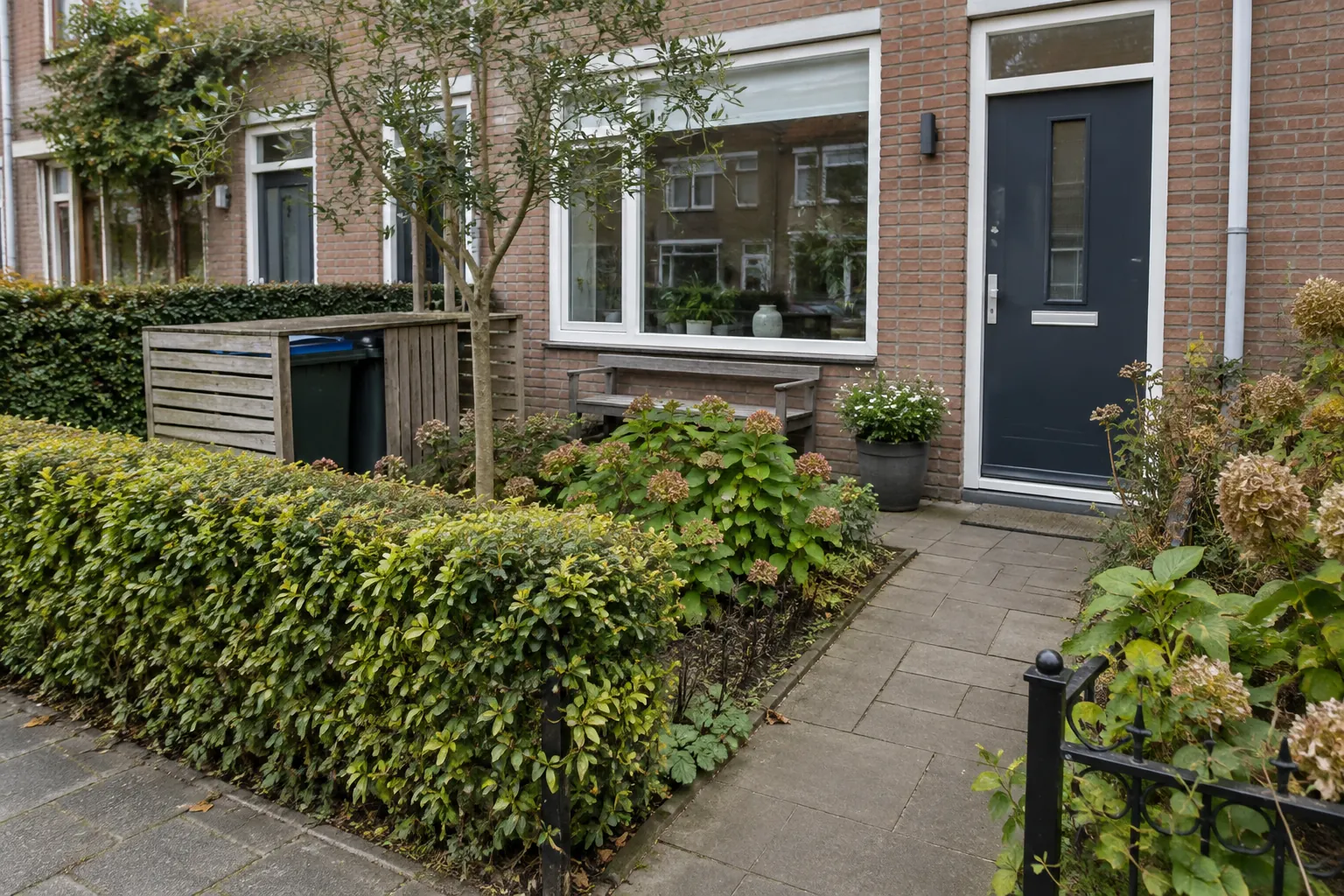 Voortuin van een rijtjeshuis met een lage haag, een houten ombouw om de containers, een smalle boom en een bestraat pad naar de voordeur — illustratief beeld